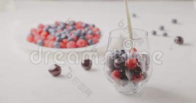 牛奶与细流倒入一个玻璃成熟浆果-樱桃，覆盆子，蓝莓在白色的桌子和一个